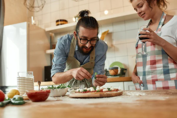 Mejor cocinero. Pareja joven haciendo pizza juntos en casa. Hombre en delantal, cocinero profesional añadiendo albahaca a la masa mientras la mujer bebe vino. Hobby, estilo de vida — Foto de Stock