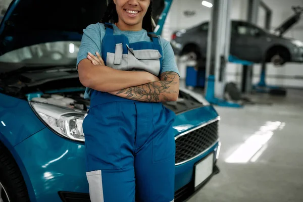 Dale un esmalte. Recortado disparo de la joven afroamericana mujer, mecánico femenino profesional sonriendo a la cámara, apoyándose en un coche en taller de reparación de automóviles — Foto de Stock