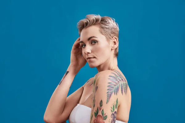 Retrato de belleza de una joven atractiva mujer medio desnuda tatuada con la piel perfecta mirando a la cámara, sosteniendo la mano cerca de su cabeza aislada sobre el fondo azul — Foto de Stock