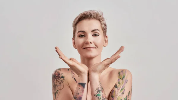 Ritratto di donna mezza nuda tatuata con i capelli corti che guarda la macchina fotografica, tenendo le palme aperte sotto il mento isolato su sfondo chiaro — Foto Stock