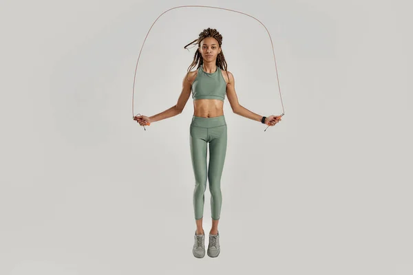 Salta todos los días. Tiro completo de mujer joven de raza mixta deportiva en ropa deportiva haciendo ejercicios con cuerda de salto aislada sobre fondo gris — Foto de Stock