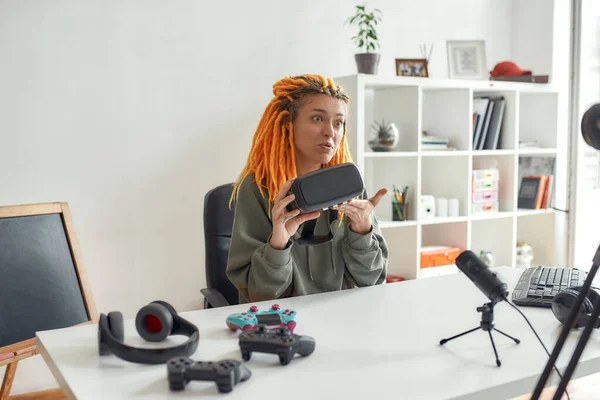 Blogueur de technologie féminine avec dreadlocks faire des visages, tenant des lunettes vr tout en enregistrant l'examen vidéo de nouveaux gadgets en utilisant un microphone à la maison — Photo