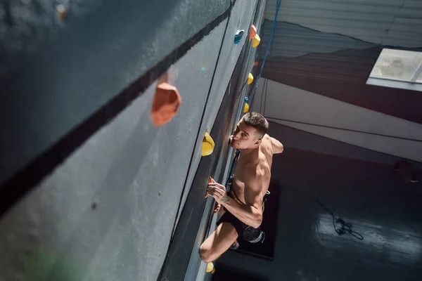 Sólo en Top. Hombre joven en equipo de seguridad y arnés mirando a la cámara mientras entrena en la pared de escalada artificial en el interior —  Fotos de Stock