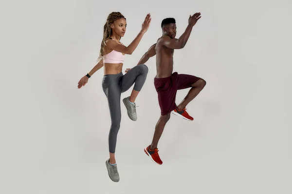 Volledige shot van jonge gespierde Afro-Amerikaanse man en sportieve gemengde ras vrouw kijken gericht tijdens het springen, oefenen geïsoleerd over grijze achtergrond. Sport en workout concept — Stockfoto