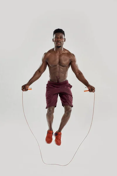 Volledige opname van jonge gespierde Afro-Amerikaanse man die naar de camera kijkt, terwijl hij oefeningen doet met springtouw geïsoleerd over een grijze achtergrond. Sport, workout, bodybuilding concept — Stockfoto