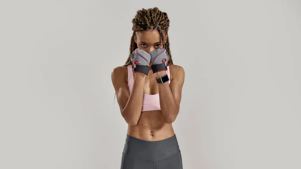 Portret van jonge sportieve gemengde ras vrouw in sportkleding kijken naar de camera, het dragen van fitness handschoenen terwijl poseren geïsoleerd over grijze achtergrond. Gezond leefstijl- en sportconcept — Stockfoto