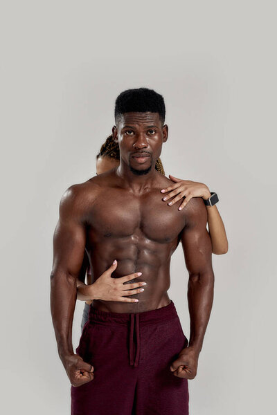 Мускулистый африканский американец смотрит в камеру, в то время как спортивная смешанная расовая женщина обнимается, прикасаясь к его телу изолированно на сером фоне. Спорт, тренировки, бодибилдинг