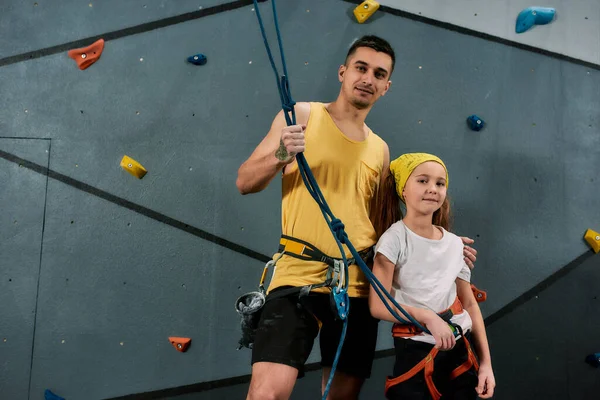 Jovem instrutor masculino e menina ativa em equipamentos de segurança olhando para a câmera, de pé contra a parede de escalada de treinamento artificial. Conceito de vida esportiva e escalada — Fotografia de Stock