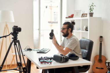 Erkek teknoloji blogcusu beklemede, kulaklıklara işaret ederek video blogu veya video blogu kaydederken ev stüdyolarındaki yeni cihazlar hakkında