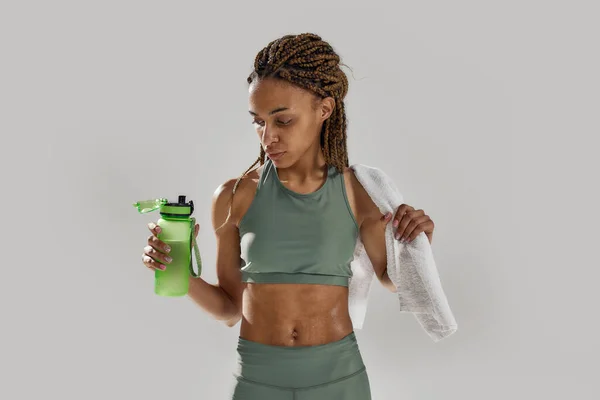 Genç sportif melez kadın, gri arka planda izole edilmiş yoğun eğitimden sonra protein içeceği şişesine bakıyor. — Stok fotoğraf