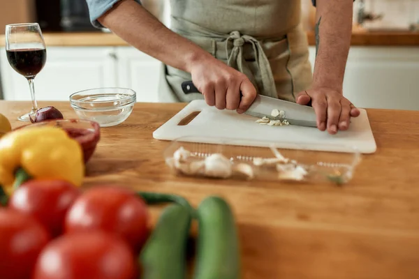 Közelkép férfi kötény peeling, vágás fokhagyma, miközben vacsorát készít, áll a konyhában. Főzés otthon koncepció — Stock Fotó