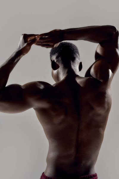 Крупный план спины молодого мускулистого африканского бодибилдера, поднимающего руки, позируя без рубашки на сером фоне. Спорт, тренировки, бодибилдинг