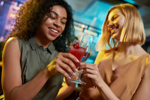 Fiesta en marcha. Atractivas mujeres jóvenes que se ven alegres mientras brindan, posando con un cóctel en sus manos. Amigos celebrando, divirtiéndose en el bar — Foto de Stock