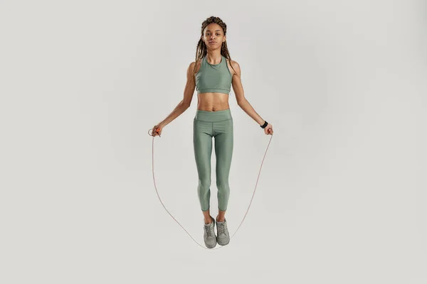 Springt. Ganzkörperaufnahme einer jungen sportlichen Mischlingsfrau in Sportbekleidung, die Übungen mit Springseil vor grauem Hintergrund macht — Stockfoto