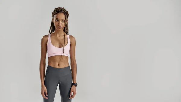 Mujer joven de raza mixta deportiva en ropa deportiva mirando a la cámara mientras posa aislada sobre un fondo gris. Estilo de vida saludable y concepto deportivo — Foto de Stock