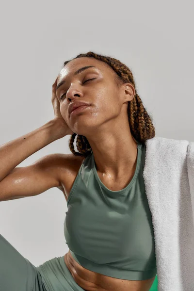 Entrenamiento productivo. Retrato de una joven mujer de raza mixta deportiva en ropa deportiva sentada en una alfombra con los ojos cerrados aislados sobre un fondo gris. Estilo de vida saludable y concepto deportivo — Foto de Stock