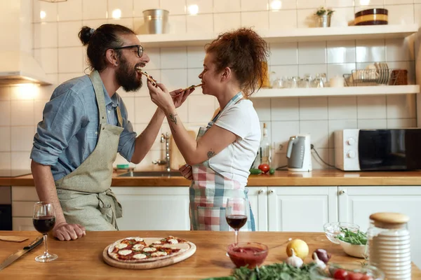 Mangia e divertiti. Giovane uomo e donna in grembiule che si nutrono la pizza appena sfornata mentre sono in cucina. Amore, concetto di relazioni — Foto Stock