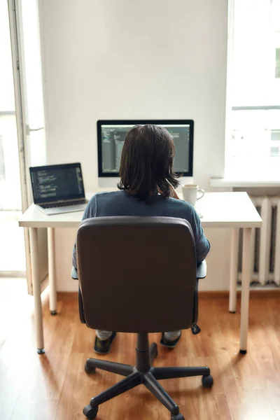 Vista posteriore di sviluppatore web maschile scrivere codice mentre seduto sul posto di lavoro e di lavoro da casa, scrivere codice. Utilizzando computer desktop e laptop. — Foto Stock