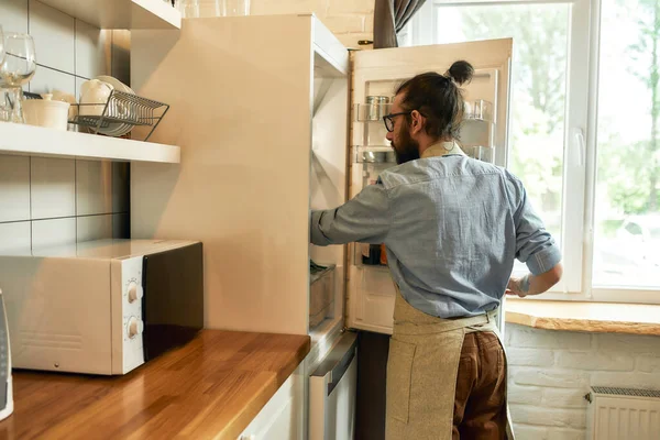 Ung man, professionell kock i förkläde ta ingredienser ur kylskåpet medan du gör dig redo att förbereda en måltid, stående i köket. Matlagning hemma koncept — Stockfoto