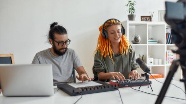 Müzik tutkusu. Rastalı kadın şarkı söylüyor ve çalıyor. Kadın ve erkek blogcu sentezleyici, davul çalar ve dizüstü bilgisayar kullanarak müzik yapıyor, video blogu veya vlog kaydediyor