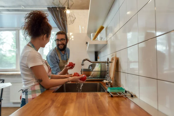 Feliz casal jovem lavar legumes, tomates na cozinha enquanto prepara os ingredientes para o jantar. Cozinhar em casa conceito — Fotografia de Stock