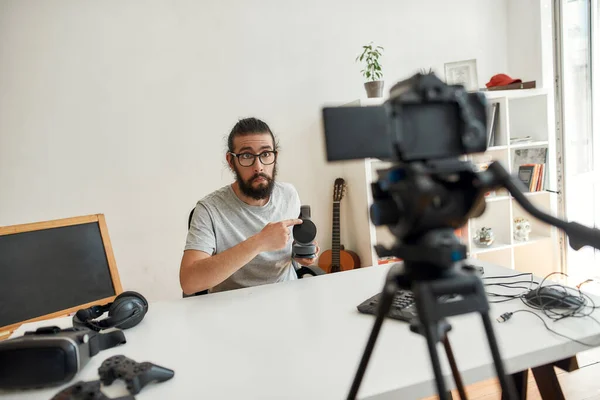 Чоловічий технологічний блогер робить обличчя, тримає, вказуючи на навушники, записує відео блог або відеоблог про нові гаджети в домашній студії — стокове фото