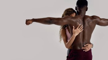 Sportif melez bir kadın. Kas yığını Afrikalı Amerikalı erkeği kucaklıyor, ona dokunuyor. Gri arka planda birlikte poz veriyorlar. Spor, spor, vücut geliştirme konsepti