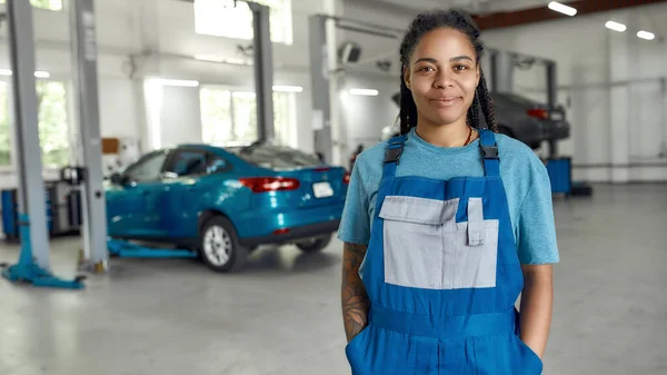 Añada nueva vida a su coche. Retrato de mujer afroamericana joven, mecánico profesional en uniforme sonriendo a la cámara, de pie en el taller de reparación de automóviles. Servicio de coches, reparación, concepto de mantenimiento — Foto de Stock