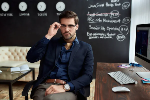 Jonge succesvolle zakenman of sales manager met een bril die naar de camera kijkt terwijl hij in het moderne kantoor zit met schoolbord op de achtergrond — Stockfoto