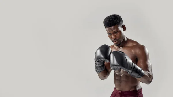 Ung muskulös afrikansk amerikansk man boxare tittar ner, bär boxningshandskar, står isolerad över grå bakgrund. Sport, träning, bodybuilding koncept — Stockfoto