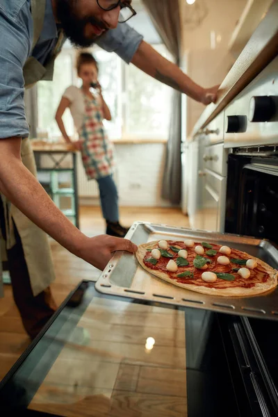 Επαγγελματίας μάγειρας φτιάχνει πίτσα στο σπίτι. Ο άνθρωπος βάζει ωμή πίτσα στο σύγχρονο φούρνο για το ψήσιμο. Γυναίκα στέκεται στο παρασκήνιο. Χόμπι, τρόπος ζωής — Φωτογραφία Αρχείου