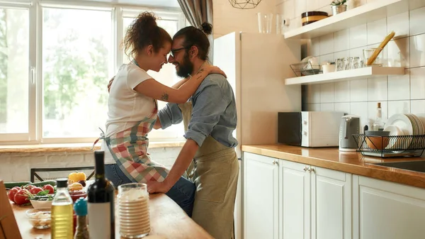 Молодий чоловік дивиться на свою дівчину, обіймаючись, стоячи на кухні. Пара обіймається під час приготування піци з овочами в приміщенні. Готуємо разом, хобі, стиль життя — стокове фото