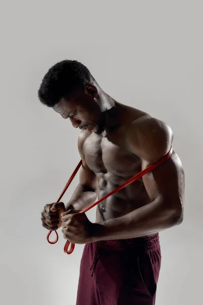 Giovane uomo afroamericano muscoloso guardando in basso, tenendo la banda di resistenza isolata su sfondo grigio. Sport, allenamento, concetto di bodybuilding — Foto Stock