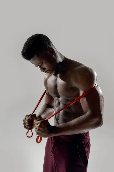 Jonge gespierde Afro-Amerikaanse man kijkt naar beneden, terwijl hij de verzetsband geïsoleerd houdt over een grijze achtergrond. Sport, workout, bodybuilding concept — Stockfoto