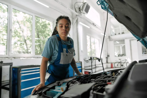 Para un mejor viaje. Mujer afroamericana joven, mecánico profesional mirando, examinando debajo de la capucha del coche en el taller de reparación de automóviles — Foto de Stock