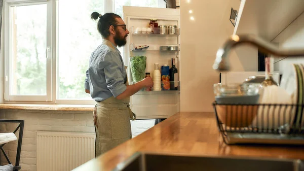 Jovem, cozinheiro profissional em avental tirando ingredientes da geladeira enquanto se prepara para preparar uma refeição, de pé na cozinha. Cozinhar em casa conceito — Fotografia de Stock