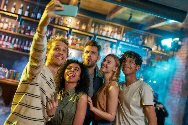 Recuerda la noche. Hombres y mujeres jóvenes sonriendo mientras toman selfie usando un teléfono inteligente. Grupo multirracial de amigos pasando el rato en la fiesta en el bar — Foto de Stock