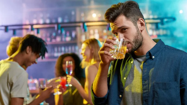 Joven guapo mirando a la cámara mientras bebe, posando con un cóctel en la mano y amigos charlando, tomando bebidas en el mostrador del bar en el fondo — Foto de Stock