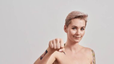 Yarı çıplak dövmeli, kısa saçlı, kameraya bakan, sıkılmış bir yumrukla el ele tutuşan, hafif arkaplanda izole edilmiş bir yumruğu yumruklayan kadın portresi.