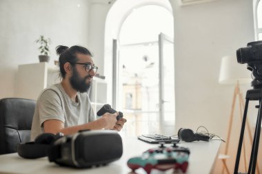 Gözlüklü bir erkek teknoloji blogcusu tatmin olmamış görünüyor, video blogu veya video blogu kaydederken oyun kumandasını elinde tutuyor.