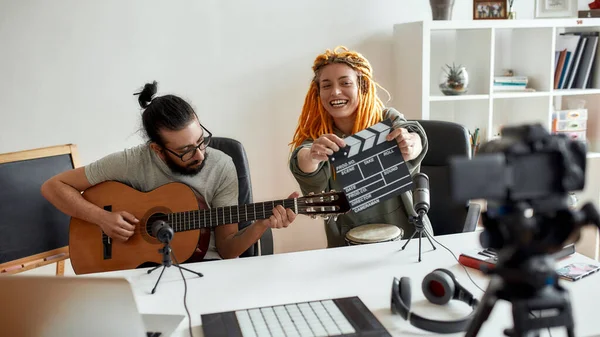 Man playing guitar and woman holding clapperboard. Couple of musicians recording video blog or vlog while making music at home — Stock Photo, Image