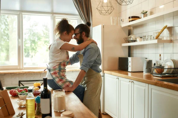 Jeune homme regardant sa petite amie, étreignant tout en se tenant dans la cuisine. Couple embrassant tout en faisant une pizza avec des légumes à l'intérieur. Cuisiner ensemble, passe-temps, mode de vie — Photo