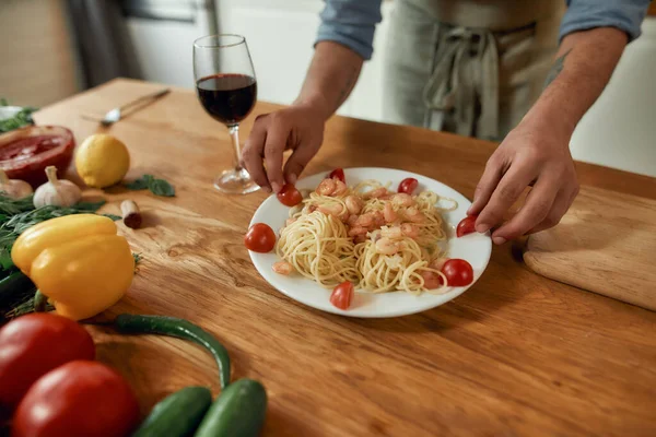Cropped shot of man, cook serving garlic butter shrimp pasta, decorating plate with tomatoes. Cooking at home, Mediterranean cuisine concept