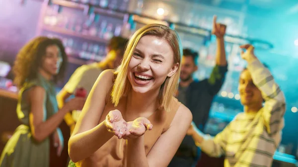 Szczęście przez całą noc. Atrakcyjna biała młoda kobieta uśmiechnięta do kamery, pozująca w nocnym klubie z konfetti w rękach. Przyjaciele tańczą, świętują, bawią się w tle — Zdjęcie stockowe