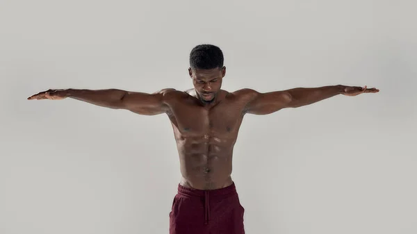 Jonge gespierde Afro-Amerikaanse man met naakte romp naar beneden kijkend terwijl hij poseerde met uitgestrekte armen geïsoleerd over grijze achtergrond. Sport, workout, bodybuilding concept — Stockfoto