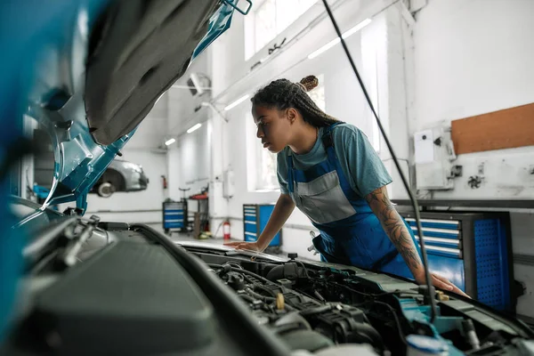 Consigue ayuda. Mujer afroamericana joven, mecánico profesional mirando, examinando debajo de la capucha del coche en el taller de reparación de automóviles — Foto de Stock