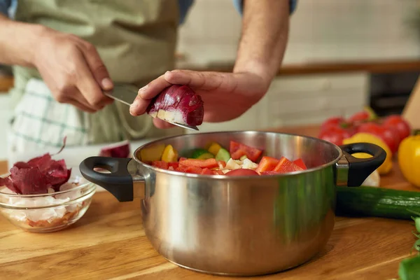 Close up of hands of man, chef cook adding onion to the pot with chopped vegetables while preparing a meal in the kitchen — Stock Photo, Image