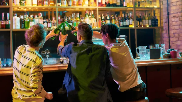 Un lugar para amigos. Tres hombres tintinean botellas de cerveza mientras están sentados en el mostrador del bar. Amigos pasando tiempo en el bar de deportes — Foto de Stock