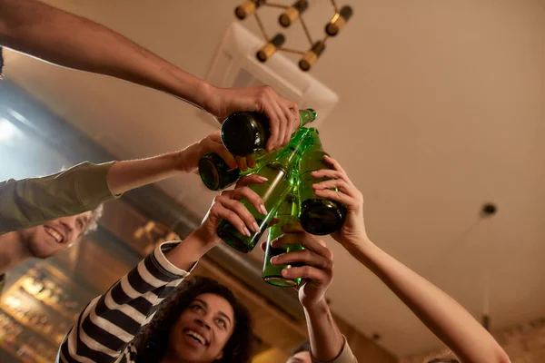 Primer plano de las manos de las personas en el bar tintineo botellas mientras beben cerveza juntos. Gente, ocio, amistad y entretenimiento — Foto de Stock