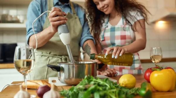 Καλλιεργημένη φωτογραφία του ανθρώπου, σεφ μαγειρεύουν χρησιμοποιώντας μπλέντερ χέρι κατά την προετοιμασία ενός γεύματος. Νεαρή γυναίκα, κοπέλα με ποδιά να ρίχνει ελαιόλαδο στην κατσαρόλα, βοηθώντας τον στην κουζίνα. — Φωτογραφία Αρχείου